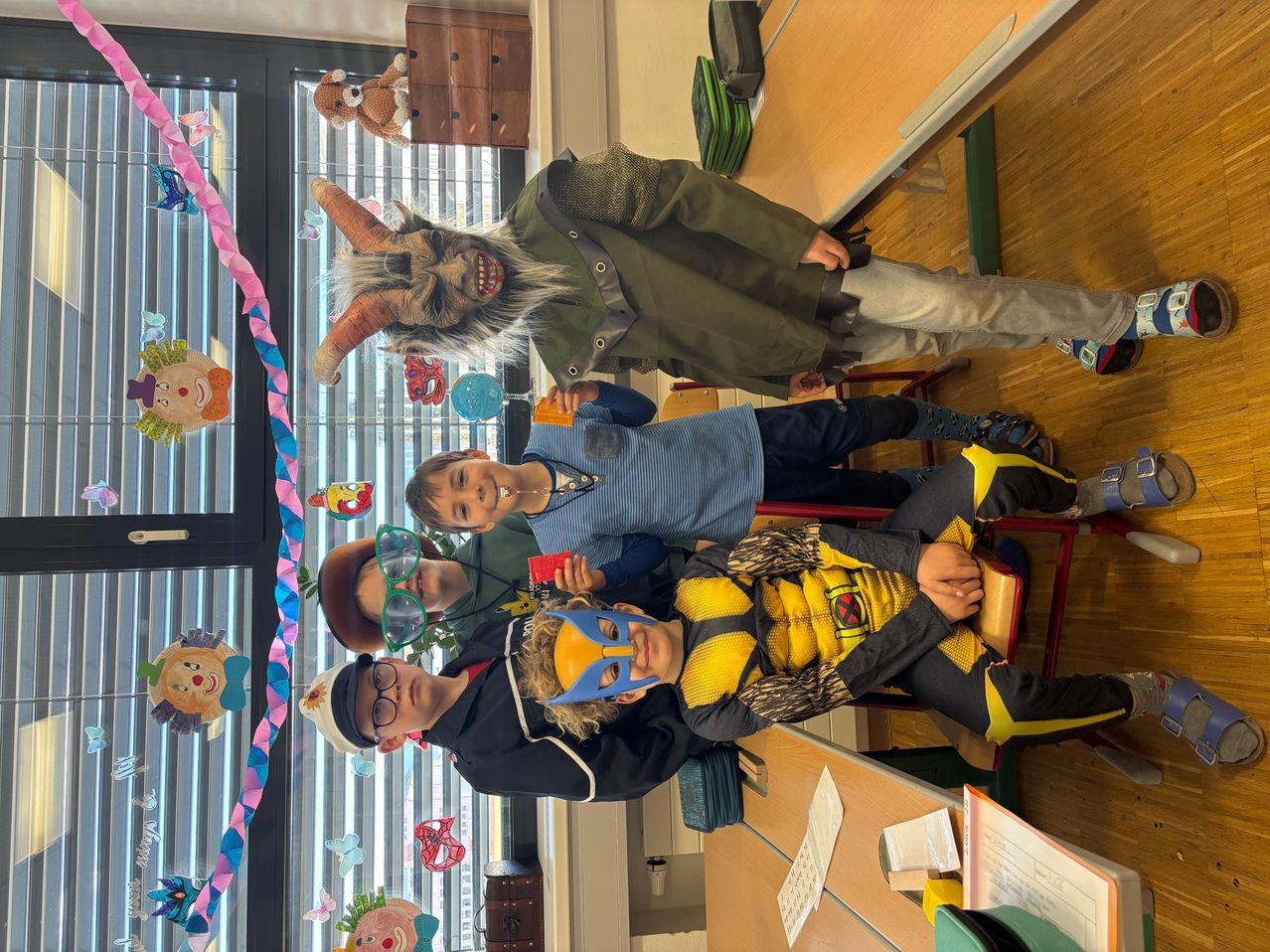 Mehrere Kinder posieren für ein Foto in einem Klassenzimmer. Eines trägt eine Maske, ein anderes hat einen Gürtel, und im Hintergrund hängen Dekorationen.