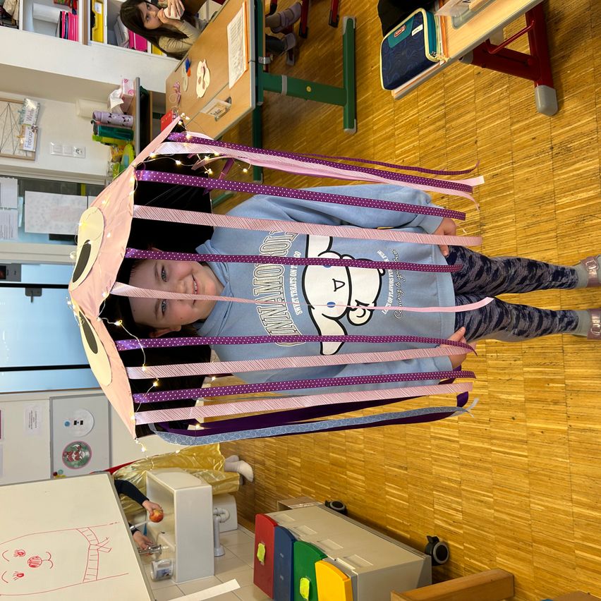 Ein junges Mädchen steht in einer Klassenzimmer in einer bunten, leuchtenden Drachenstruktur und lächelt. Der Boden ist aus Holz, und andere Kinder sind im Hintergrund zu sehen.