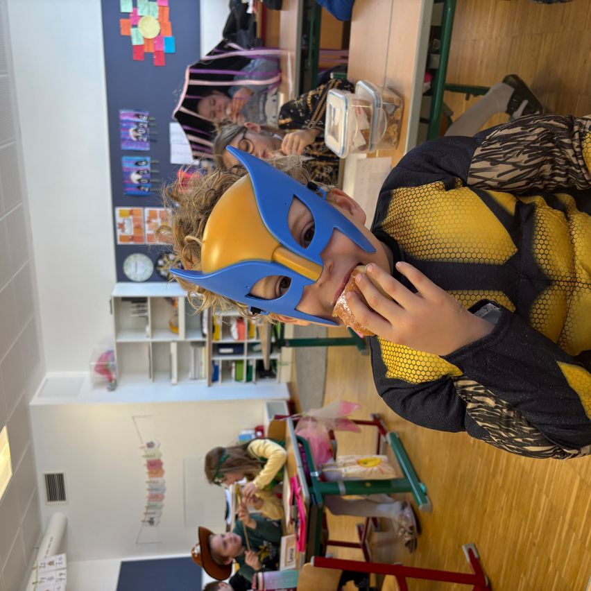 Ein junger Junge in einem Superheldenkostüm isst einen Sandwich in einem Klassenzimmer mit anderen Schülern.