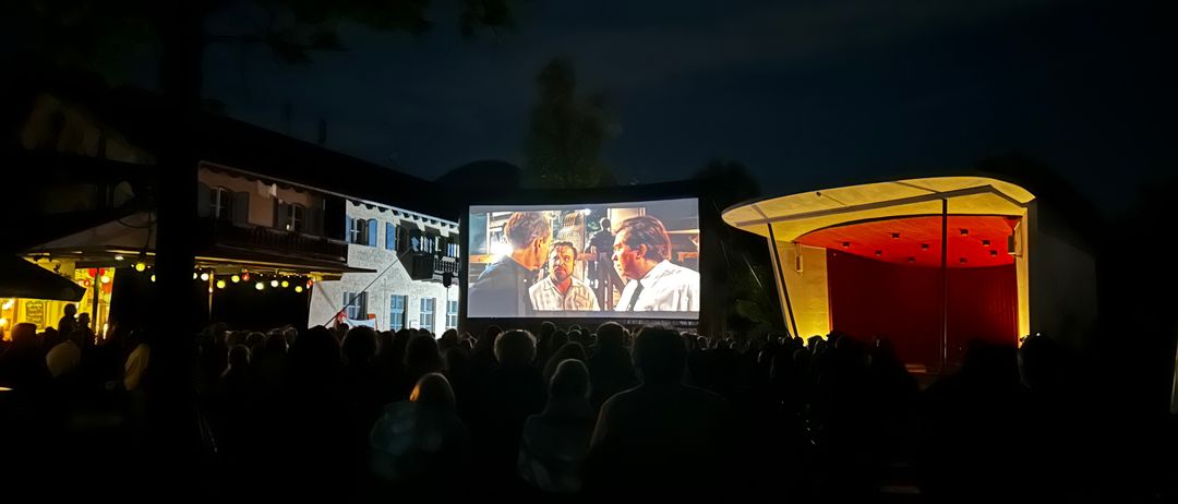 In der Nacht sieht eine große Menschenmenge einen Film auf einer Projektionsleinwand im Freien. Die Leinwand zeigt einen Mann in einem weißen Hemd und einen Mann in einem gelben Hemd.