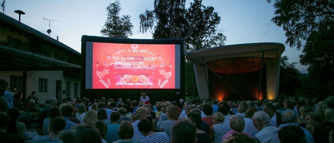 Eine Menschenmenge schaut einen Film auf einer Leinwand im Freien. Die Leinwand zeigt ein Plakat für ein Musikfestival.