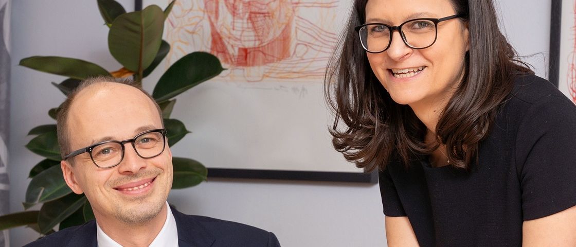 Ein Mann und eine Frau lächeln und posieren für ein Foto. Der Mann trägt einen Anzug und eine Krawatte, und die Frau trägt eine Brille und ein schwarzes Oberteil. Hinter ihnen steht eine Pflanze und ein gerahmtes Bild an der Wand.