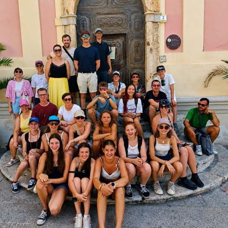 Eine Gruppe von Männern und Frauen posiert für ein Foto vor einem alten Gebäude mit einer Holztür und einer Plakette an der Wand.