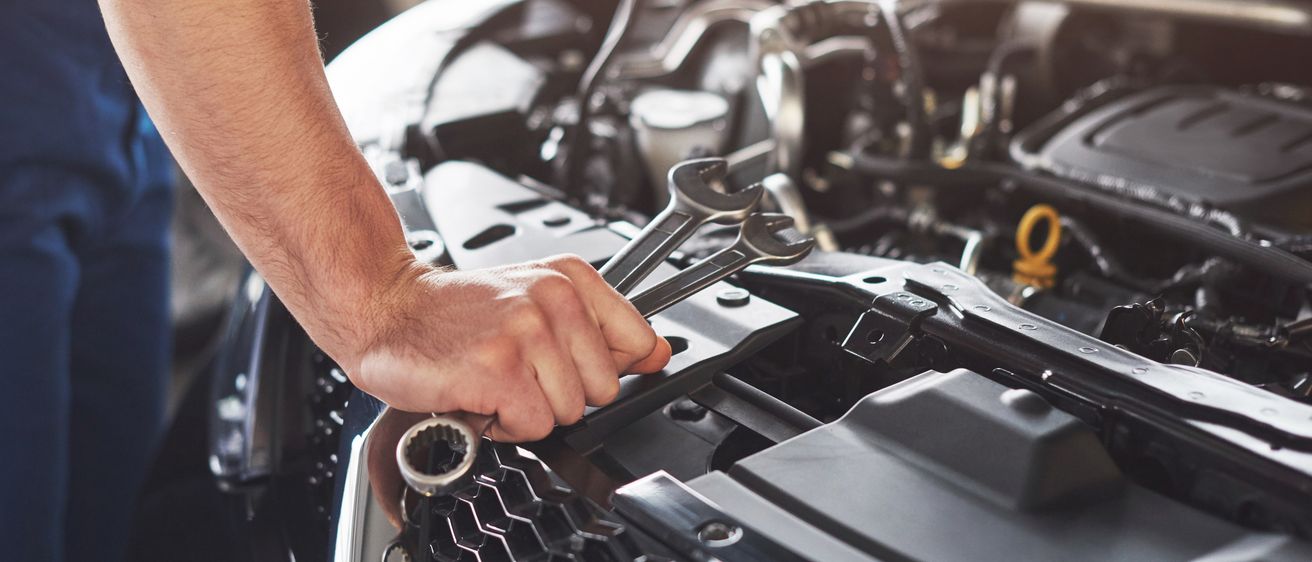 Eine Person arbeitet mit einem Schraubenschlüssel an einem Automotor in einer Garage. Die Motorhaube ist geöffnet und der Motor ist freigelegt.