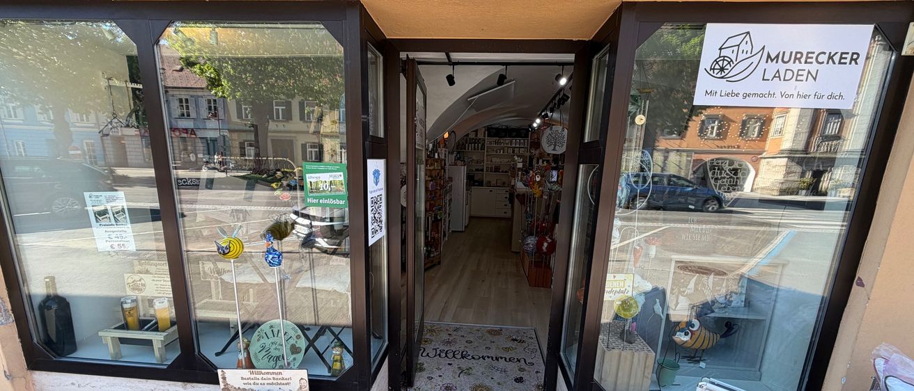 Der Eingang eines Ladens mit einem Schild, das 'Mureck Laden' steht. Der Laden hat Glastüren und Fenster, einen Willkommensmatte und einen Topf mit Pflanzen. Im Inneren gibt es Regale mit Gegenständen und einem Kühlschrank. Ein Auto ist draußen geparkt.