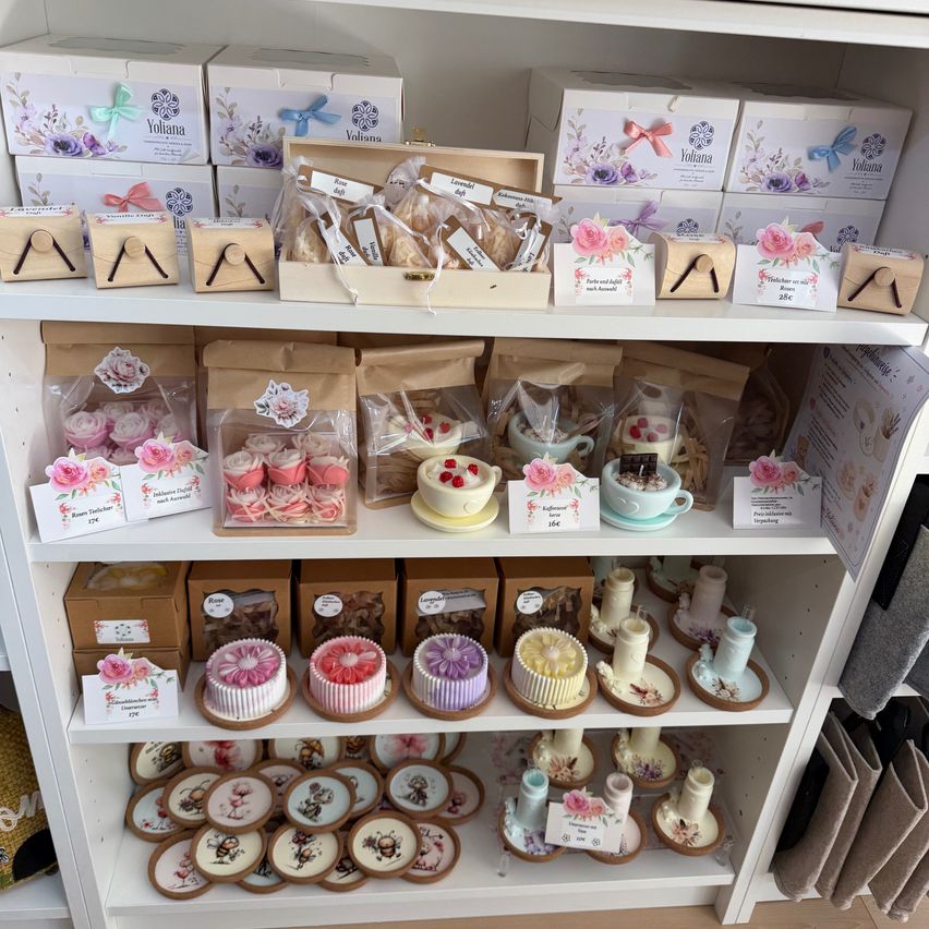 A white shelf displays an assortment of decorative items, including cakes, cupcakes, and cookies, all wrapped in festive packaging with floral designs.