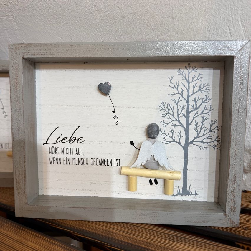 A framed artwork with a wooden bench, an angel sitting on it, a heart floating in the air, and a tree. The word 'Liebe' is written in black on a white background.