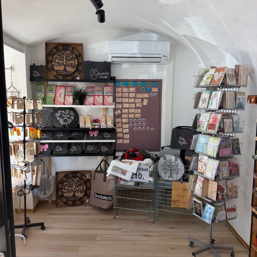 A boutique interior with wooden shelves, a clock, and various items for sale. The shelves are filled with bags, cards, and decorative items. An air conditioner is mounted on the wall.