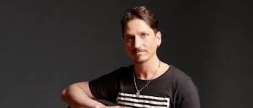 Ein Mann in einem schwarzen T-Shirt und Jeans sitzt auf einem Hocker und spielt Gitarre.