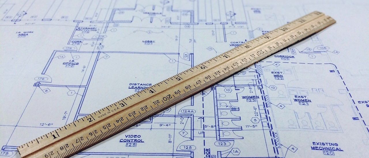 Eine Holzlineal liegt auf einer Blaupause mit einem Grundriss, der Räume mit den Bezeichnungen 'MÄNNER', 'FRAUEN', 'KONFERENZ' und 'VIDEOSTEUERUNG' enthält.