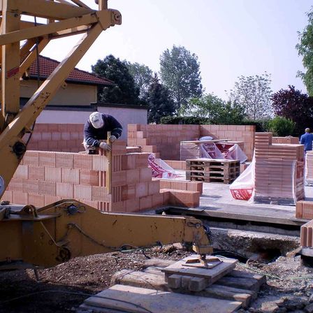 Ein Bauarbeiter in einer weißen Mütze und Handschuhen legt Ziegelsteine an einer Wand, mit einem gelben Kran in der Nähe und mehreren Stapeln von Ziegelsteinen auf dem Boden.