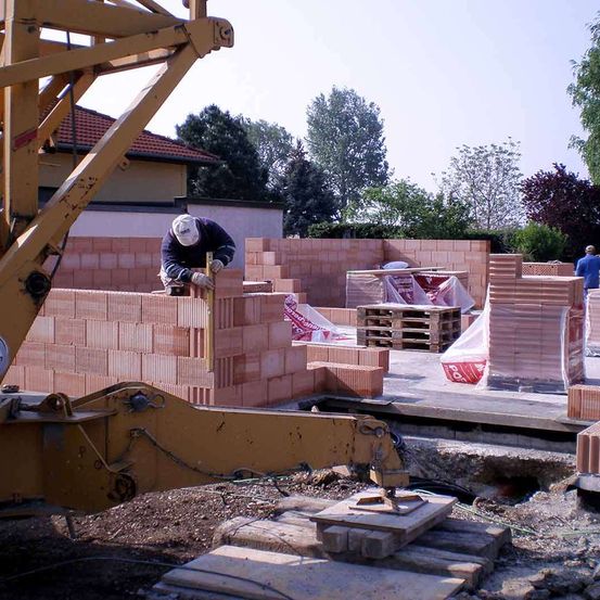 Ein Bauarbeiter in einer weißen Mütze und Handschuhen legt Ziegelsteine an einer Wand, mit einem gelben Kran in der Nähe und mehreren Stapeln von Ziegelsteinen auf dem Boden.