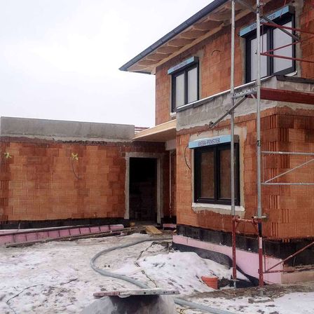 Ein Haus im Bau mit Ziegelwänden und Gerüsten wird gezeigt. Es hat ein Glasfenster mit einem blauen Schild, auf dem 'Weba-Fenster' steht.