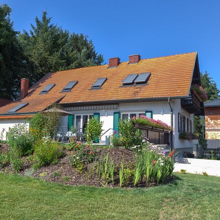 Ein weißes Haus mit einem braunen Dach und Solarpaneelen ist von einem Garten umgeben. Der Garten zeichnet sich durch eine Vielfalt an Pflanzen und Blumen aus, einige davon in Töpfen.