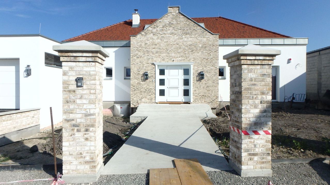Ein modernes Backsteinhaus mit rotem Dach, weißer Tür und zwei Backsteinsäulen auf jeder Seite. Das Haus ist im Bau mit einem Betonweg.