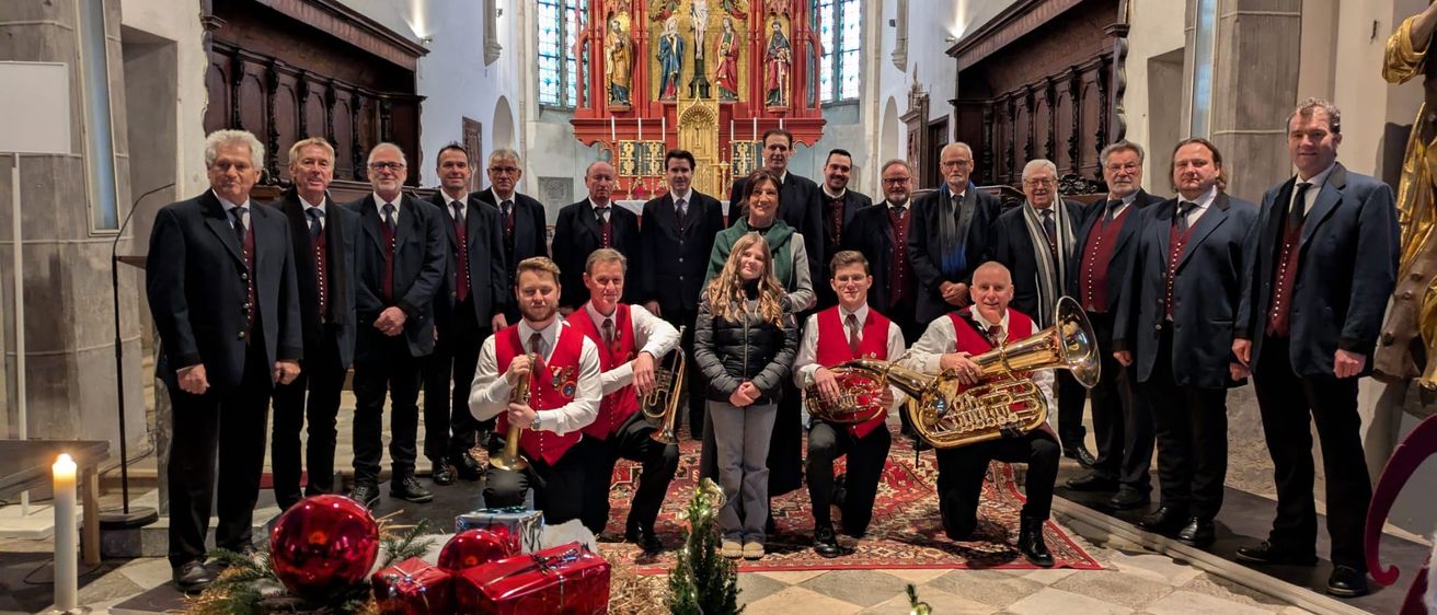 Eine Gruppe von Menschen in formeller Kleidung posiert für ein Foto in einer Kirche mit Weihnachtsdekoration. Einige halten Blechblasinstrumente. Hinter ihnen ist ein geschmückter Altar mit Statuen zu sehen.