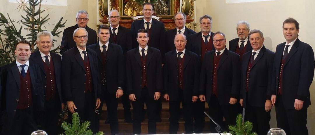 Ein Männerchor in schwarzen Anzügen und Westen steht vor einem geschmückten Altar, mit einem religiösen Gemälde im Hintergrund.