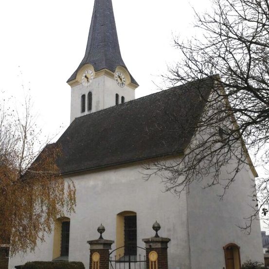 Eine kleine Kirche mit einem Turm und zwei Uhren, einem schwarzen Dach und gelben Akzenten. Sie hat ein Tor vorn und ist von Bäumen umgeben.