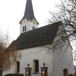 Eine kleine Kirche mit einem Turm und zwei Uhren, einem schwarzen Dach und gelben Akzenten. Sie hat ein Tor vorn und ist von Bäumen umgeben.