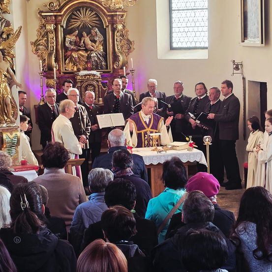 Ein Priester leitet einen Gottesdienst in einer Kirche. Der Altar ist mit Blumen und Kerzen geschmückt. Chormitglieder stehen hinter dem Priester und singen. Die Gemeinde sitzt aufmerksam.