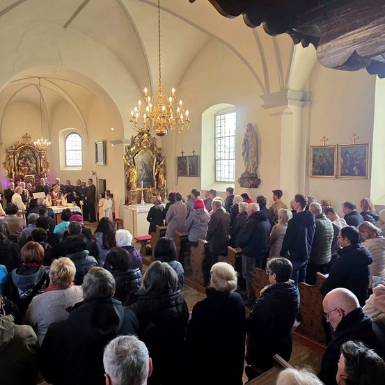 Eine große Gruppe von Menschen in einer Kirche, einige mit Hüten, beten und stehen auf Kirchenbänken mit religiösen Symbolen an den Wänden.