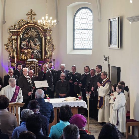 Ein Gottesdienst mit Priester, Chor und Gemeinde. Ein goldener Altar mit religiösen Bildern ist vorhanden.