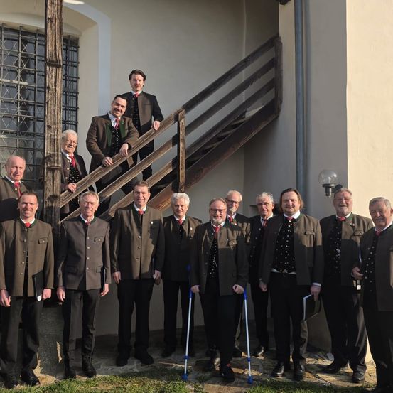 Eine Gruppe von Männern in traditioneller Kleidung steht vor einem Gebäude mit einer Treppe und einer Lampe. Einige halten Stöcke.