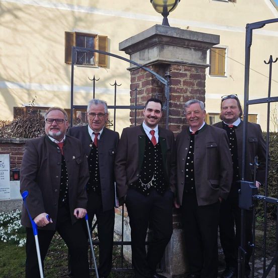 Fünf Männer in traditioneller Kleidung stehen vor einem Gebäude und lächeln für ein Foto. Hinter ihnen befindet sich ein Ziegelsäule mit einer Laterne und einem geschlossenen Tor.