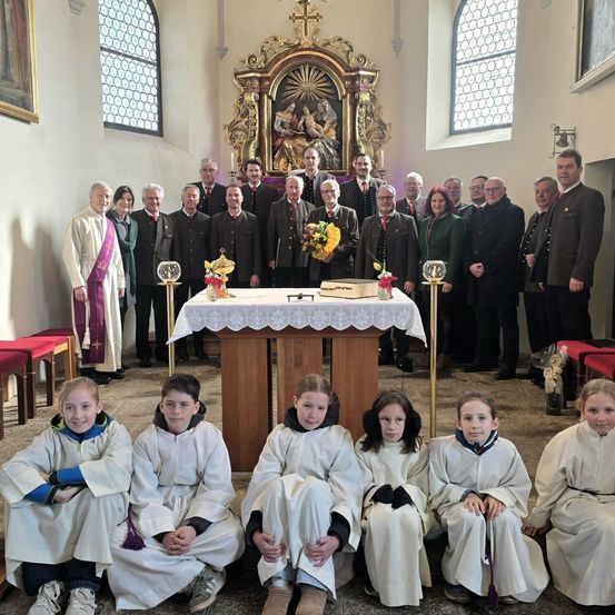 Eine Gruppe von Menschen steht und sitzt in einer Kirche, gekleidet in weiße Gewänder. Ein Priester steht an einem Tisch mit Blumen. Dahinter stehen andere in Anzügen und Krawatten vor einem Altar.