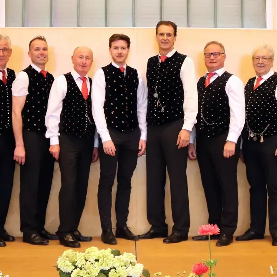 Sieben Männer in passenden Westen und Krawatten posieren für ein Foto. Sie stehen in einer Reihe und lächeln. Vor ihnen stehen Blumen.