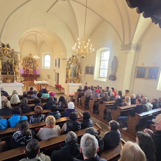 Eine große Gemeinde versammelt sich in einer Kirche mit hohen Decken und Holzbänken. Der Altar im Vordergrund ist mit religiöser Kunst geschmückt und eine Kronleuchter hängt über dem Raum. Die Menschen sitzen aufmerksam, einige in Winterkleidung.