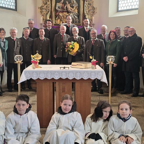 Eine Gruppe von Menschen steht hinter einem Altar in einer Kirche, geschmückt mit Blumen und Kerzen. Männer in Anzügen und Frauen in Mänteln stehen hinter Kindern in weißen Gewändern.