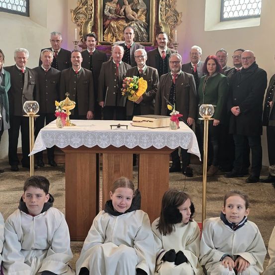Eine Gruppe von Erwachsenen und Kindern in weißen Gewändern posiert vor einem Altar mit Blumen, Kerzen und einer Box. Sie befinden sich in einer Kirche.
