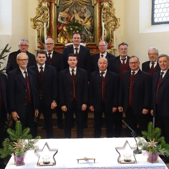 Eine Gruppe formell gekleideter Männer steht vor einem Altar und lächelt für ein Foto. Dahinter ist ein kunstvolles Gemälde und Kerzen zu sehen.