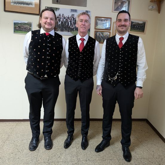 Drei Männer stehen in einem Raum zusammen, jeder trägt schwarze Westen und Hosen. Sie lächeln und scheinen für ein Foto zu posieren. Hinter ihnen hängen verschiedene gerahmte Bilder an der Wand.