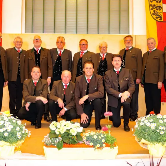 Eine Gruppe von Männern in passenden braunen Anzügen und roten Krawatten posiert für ein Foto auf einer Bühne mit Blumen.