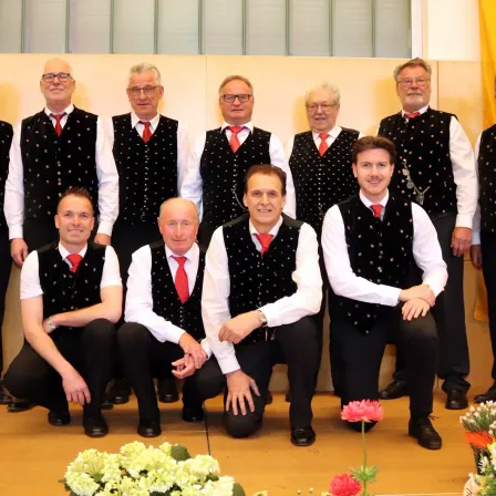 Eine Gruppe älterer Männer in schwarzen und weißen Westen mit roten Krawatten, die stehen und knien, lächeln und für ein Foto in einem Raum mit Holzboden und Blumen posieren.