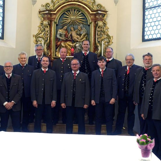 Eine Gruppe formell gekleideter Männer steht vor einem kunstvollen Altar und lächelt für ein Foto. Der Altar zeigt ein religiöses Bild und Kerzen.