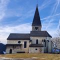 Eine Kirche mit einem Kirchturm steht auf einem Hügel, umgeben von Bäumen. Der Himmel ist blau mit weißen Wolken, und der Boden ist schneebedeckt.
