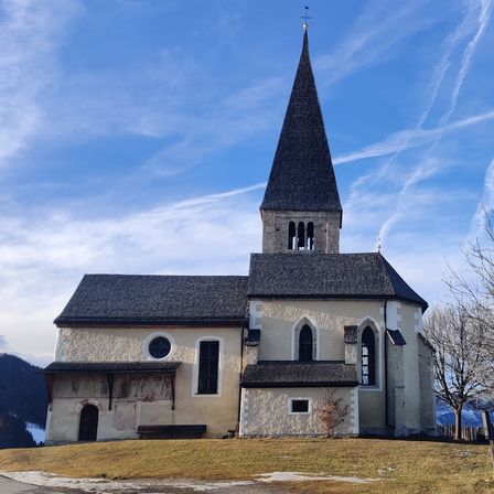 Eine Kirche mit einem Kirchturm steht auf einem Hügel, umgeben von Bäumen. Der Himmel ist blau mit weißen Wolken, und der Boden ist schneebedeckt.