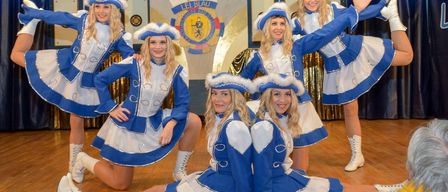 Fünf Frauen in blauen und weißen Kostümen mit Hüten posieren für ein Foto auf einer Bühne mit einem großen Logo im Hintergrund.