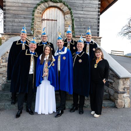 Eine Gruppe von Personen in zeremonieller Kleidung steht vor einem Gebäude, eine Frau in der Mitte hält eine Trophäe.