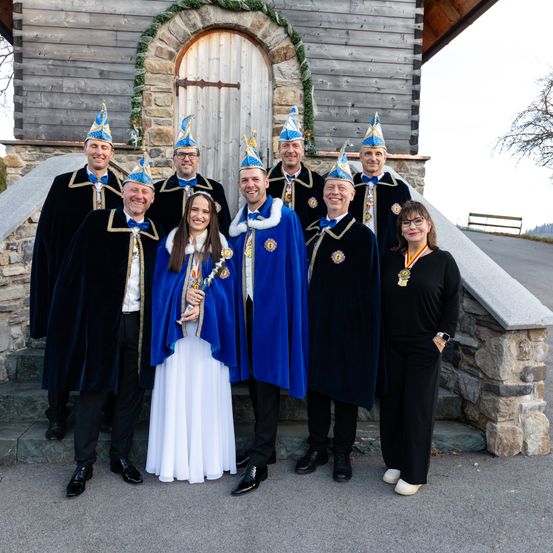 Eine Gruppe von Personen in zeremonieller Kleidung steht vor einem Gebäude, eine Frau in der Mitte hält eine Trophäe.
