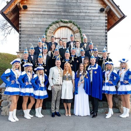Eine Gruppe von Menschen in traditionellen Kostümen steht vor einem hölzernen Gebäude mit Steindetails und lächelt für ein Foto.