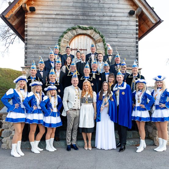 Eine Gruppe von Menschen in traditionellen Kostümen steht vor einem hölzernen Gebäude mit Steindetails und lächelt für ein Foto.