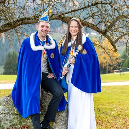 Ein Mann und eine Frau in traditioneller Kleidung posieren unter einem Baum und lächeln. Der Mann hält einen Zepter und beide tragen Medaillen. Im Hintergrund sind Berge und grüner Rasen zu sehen.