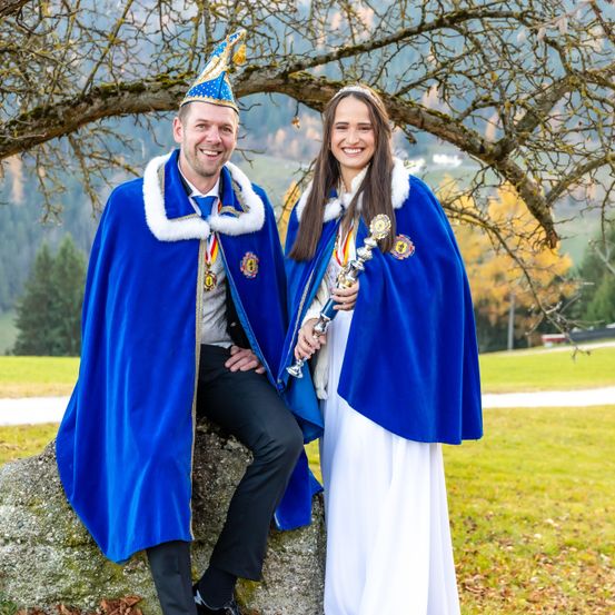 Ein Mann und eine Frau in traditioneller Kleidung posieren unter einem Baum und lächeln. Der Mann hält einen Zepter und beide tragen Medaillen. Im Hintergrund sind Berge und grüner Rasen zu sehen.