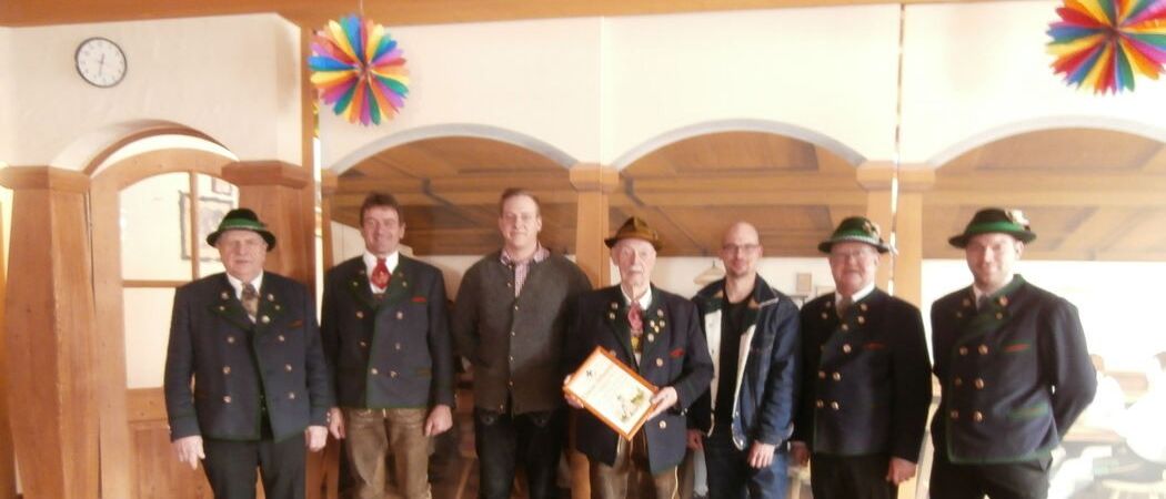 Eine Gruppe von Männern in traditioneller Kleidung posiert für ein Foto. Ein Mann hält ein gerahmtes Zertifikat. Sie stehen in einem Raum mit Holzbalken und einer festlichen Dekoration.