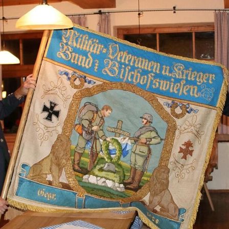 Zwei Personen halten eine Fahne mit einer Illustration von zwei Soldaten, einer mit Gewehr und der andere legt einen Kranz. Die Fahne hat goldene Verzierungen und deutschen Text.