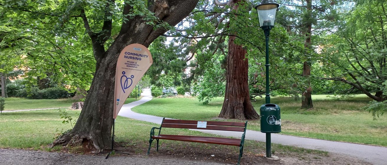 Eine Parkbank unter einem Baum mit einem Banner für Gemeindekrankenpflege. Ein Lampenpfosten und ein Mülleimer sind in der Nähe.
